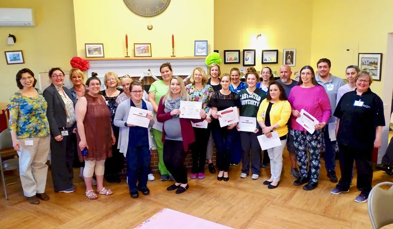 Group of people holding certificates, standing in a brightly lit room with awards, smiling, and posing for a photo.
