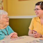 Elderly woman in blue shirt and young woman in yellow shirt laughing at a table, cards in hand.