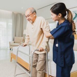 Caregiver assisting person using a walker to stand next to bed. Interior bedroom with neutral tones.
