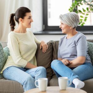 Two women seated on a couch, talking. One has a ponytail, the other gray hair. They are smiling and holding mugs.