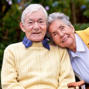 Elderly couple smiling, woman's head on man's shoulder. They are outdoors, man in yellow sweater, woman in yellow jacket.