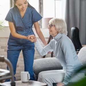 Caregiver assisting a senior woman from a chair; both smiling, in a home setting.