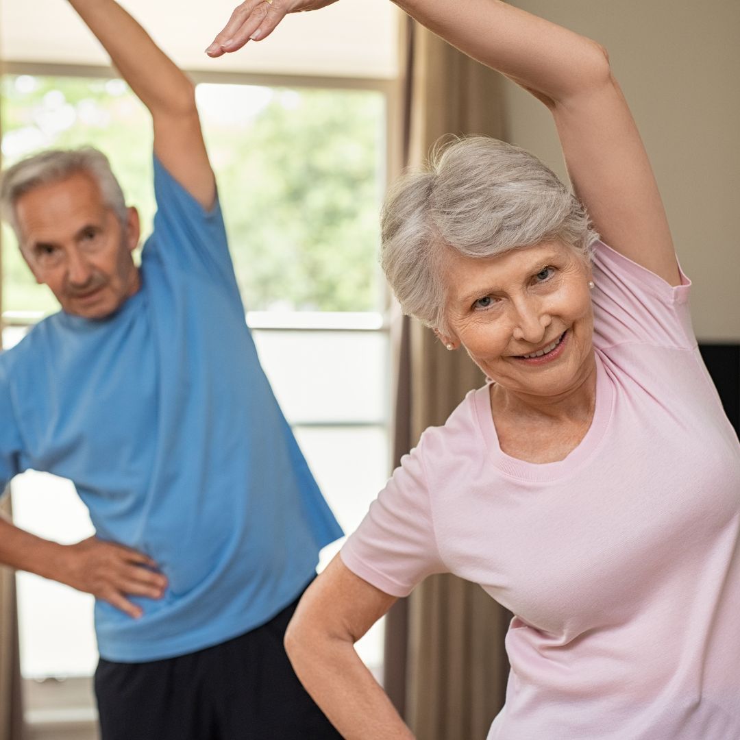 Couple stretching indoors. Man in blue shirt, woman in pink shirt, both smiling.