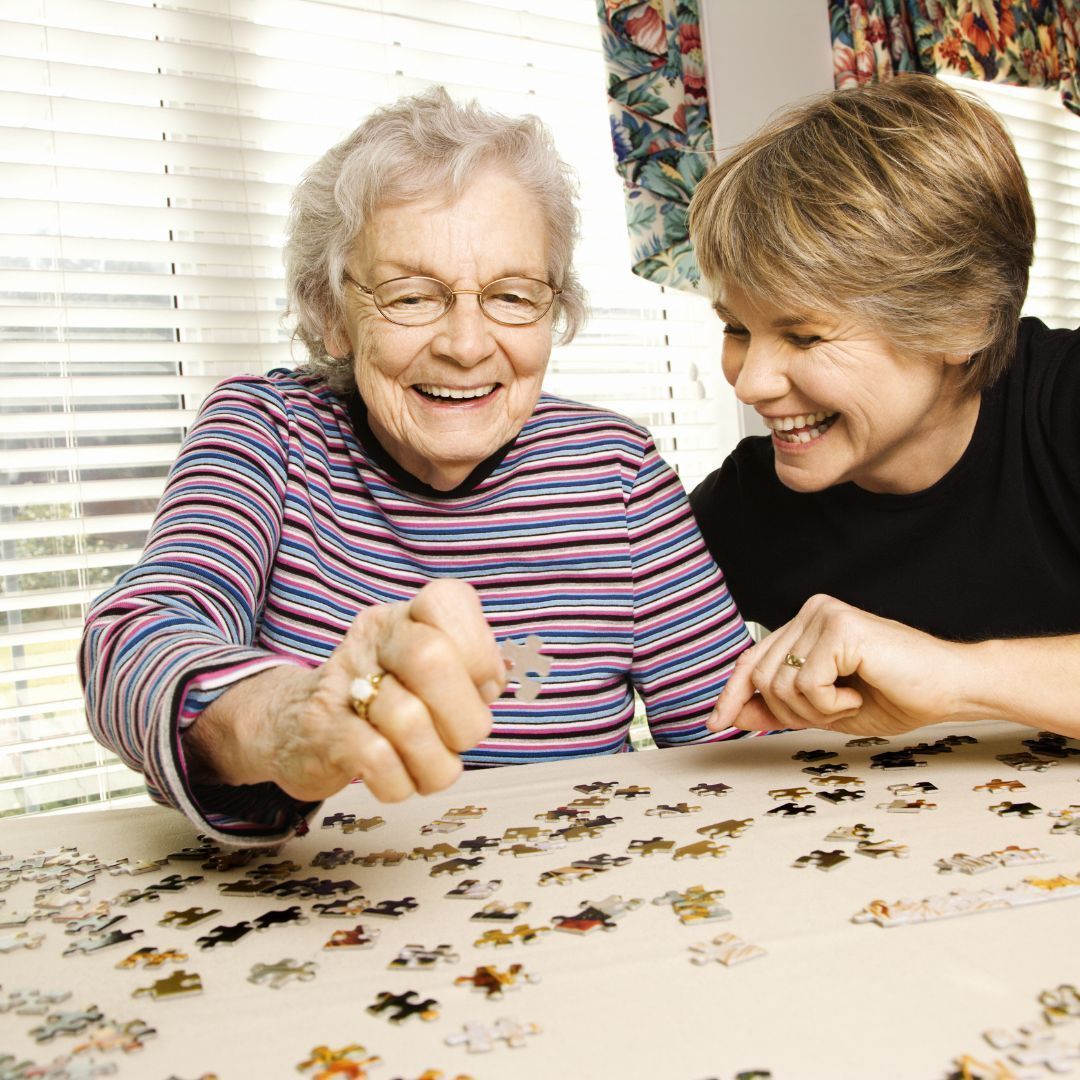 Woman and older woman smiling, working on a jigsaw puzzle at a table.