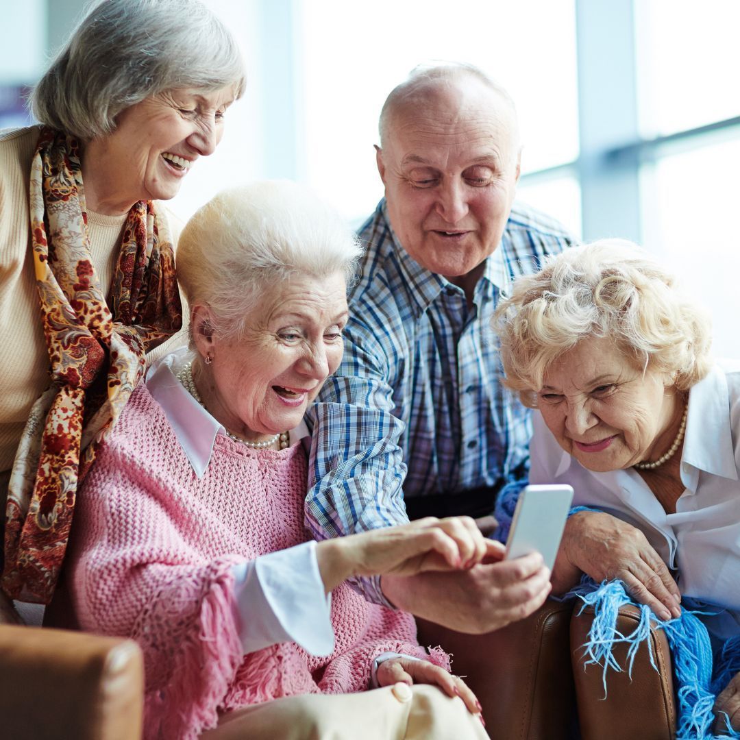 Four older adults looking at a smartphone, smiling. One holds the phone, others peer over. Indoors.