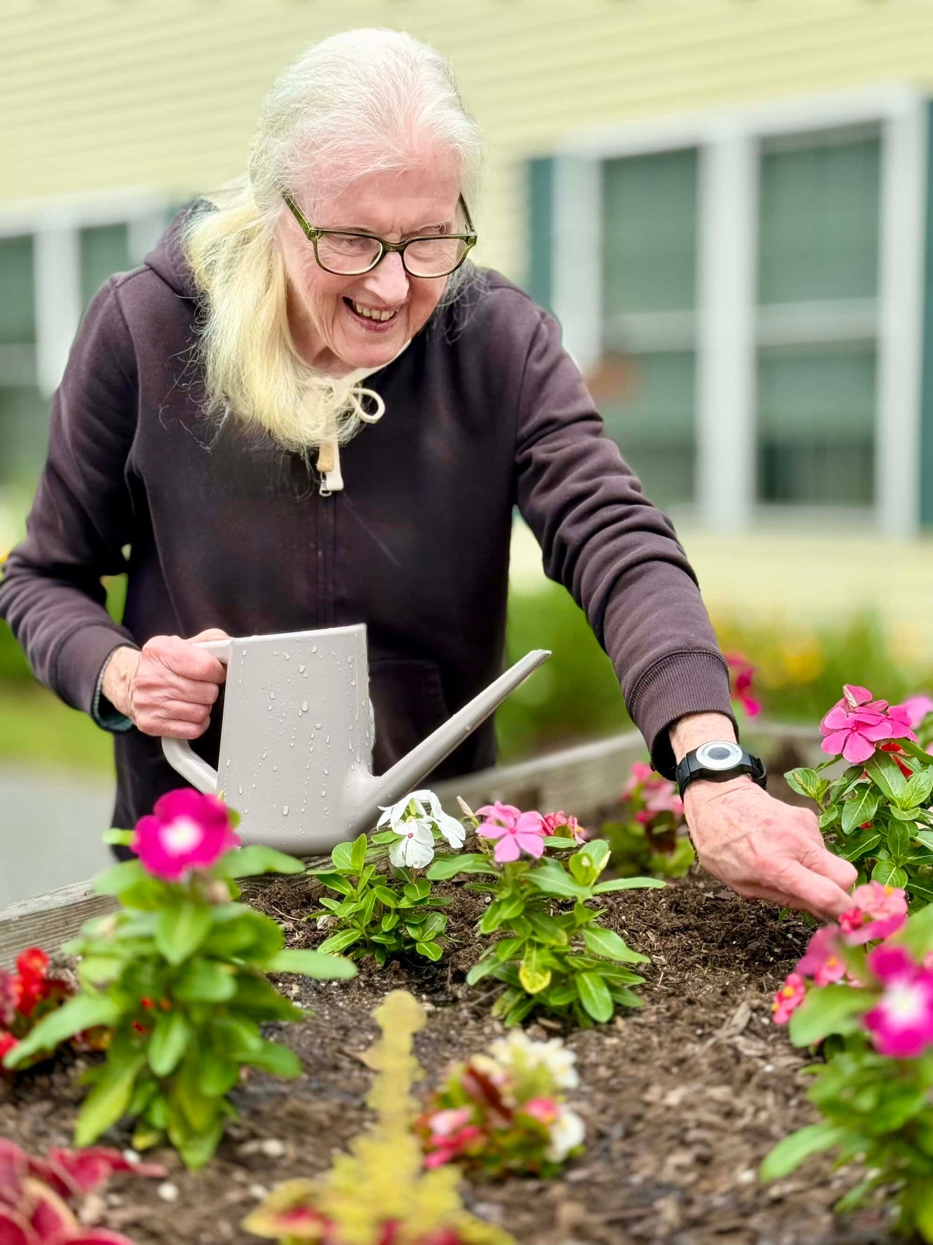 Elderly woman waters flowers in a garden bed, smiling.