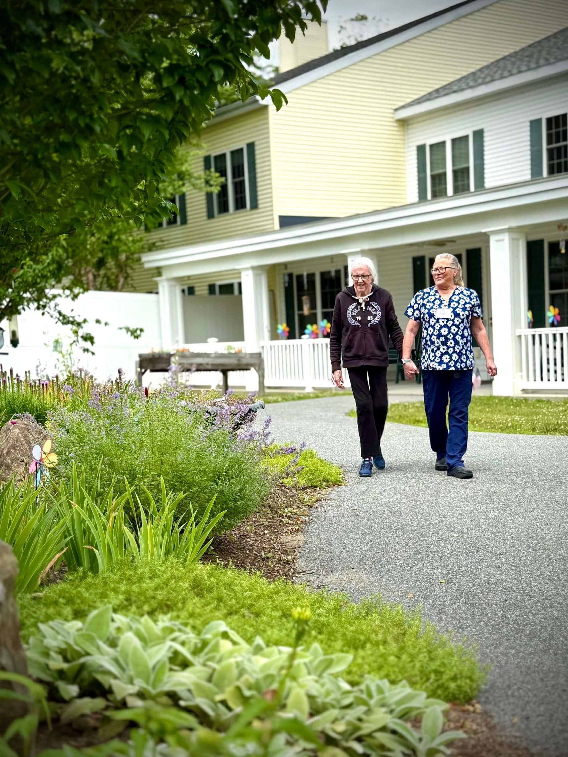 Two people walking, holding hands, on a gravel path in front of a building with flowers.