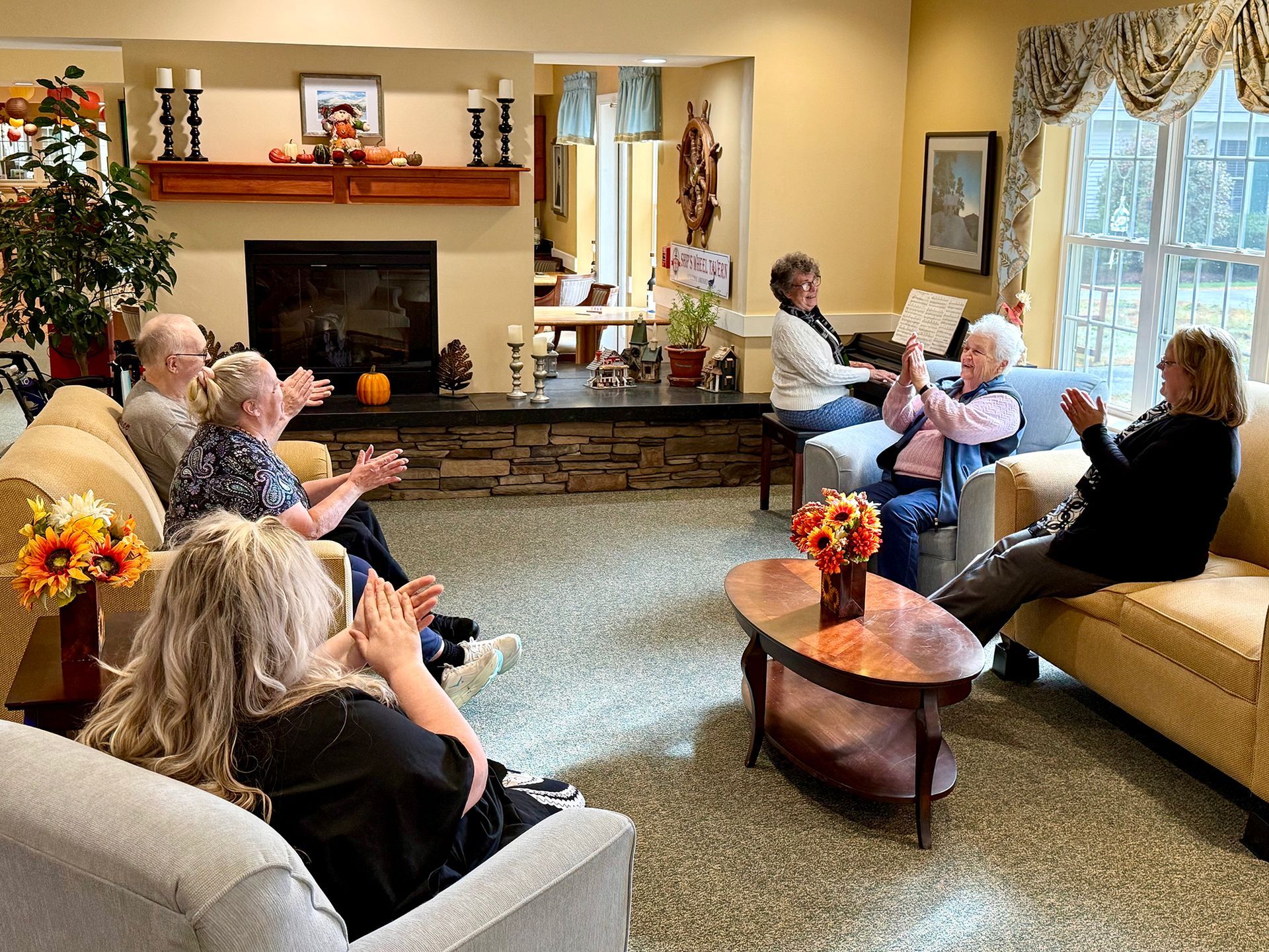 A group of people sit in a cozy lounge area, some clapping, gathered around a central coffee table near a fireplace.