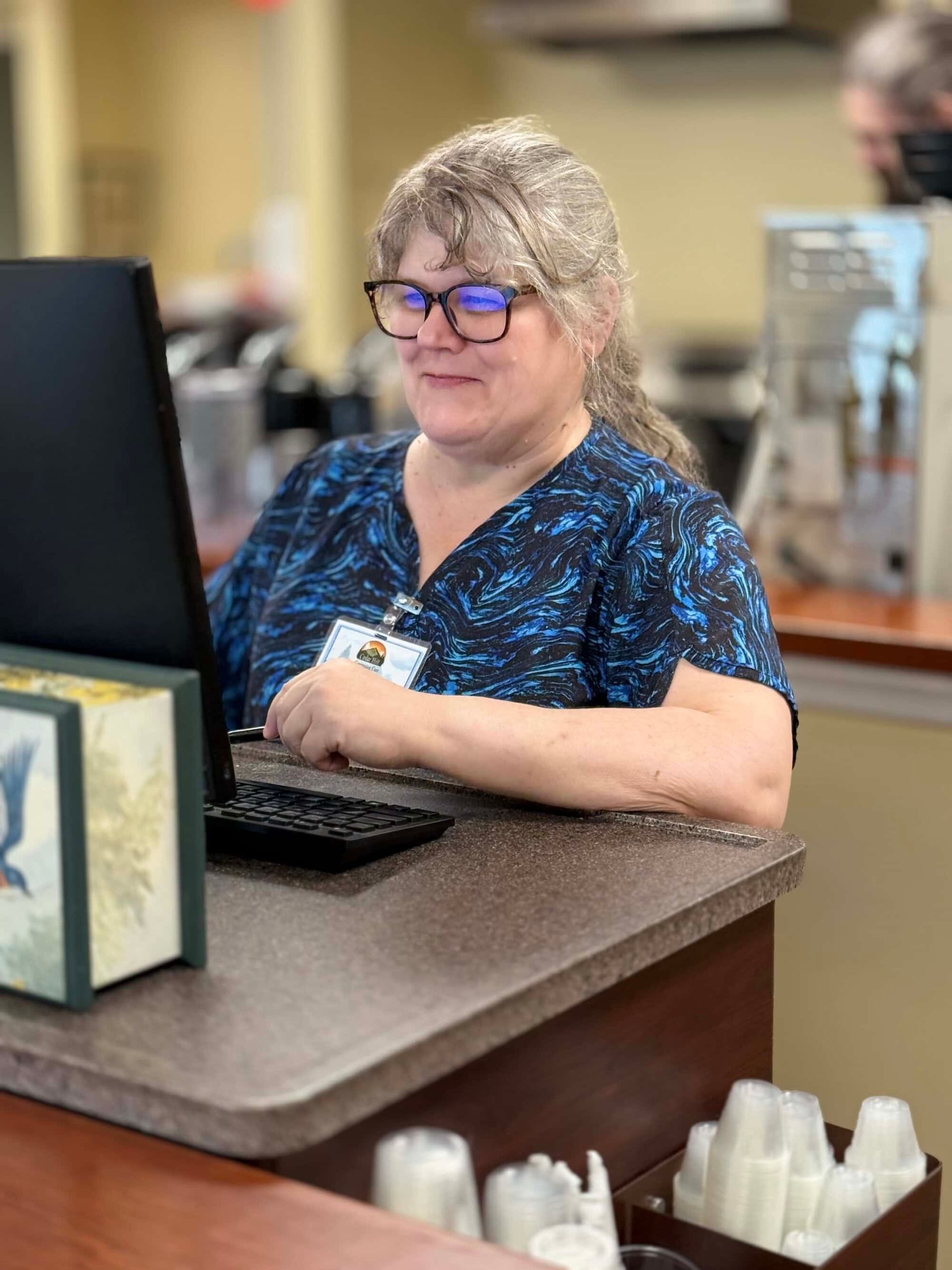 Woman wearing glasses and blue patterned top, working on a computer at a counter.