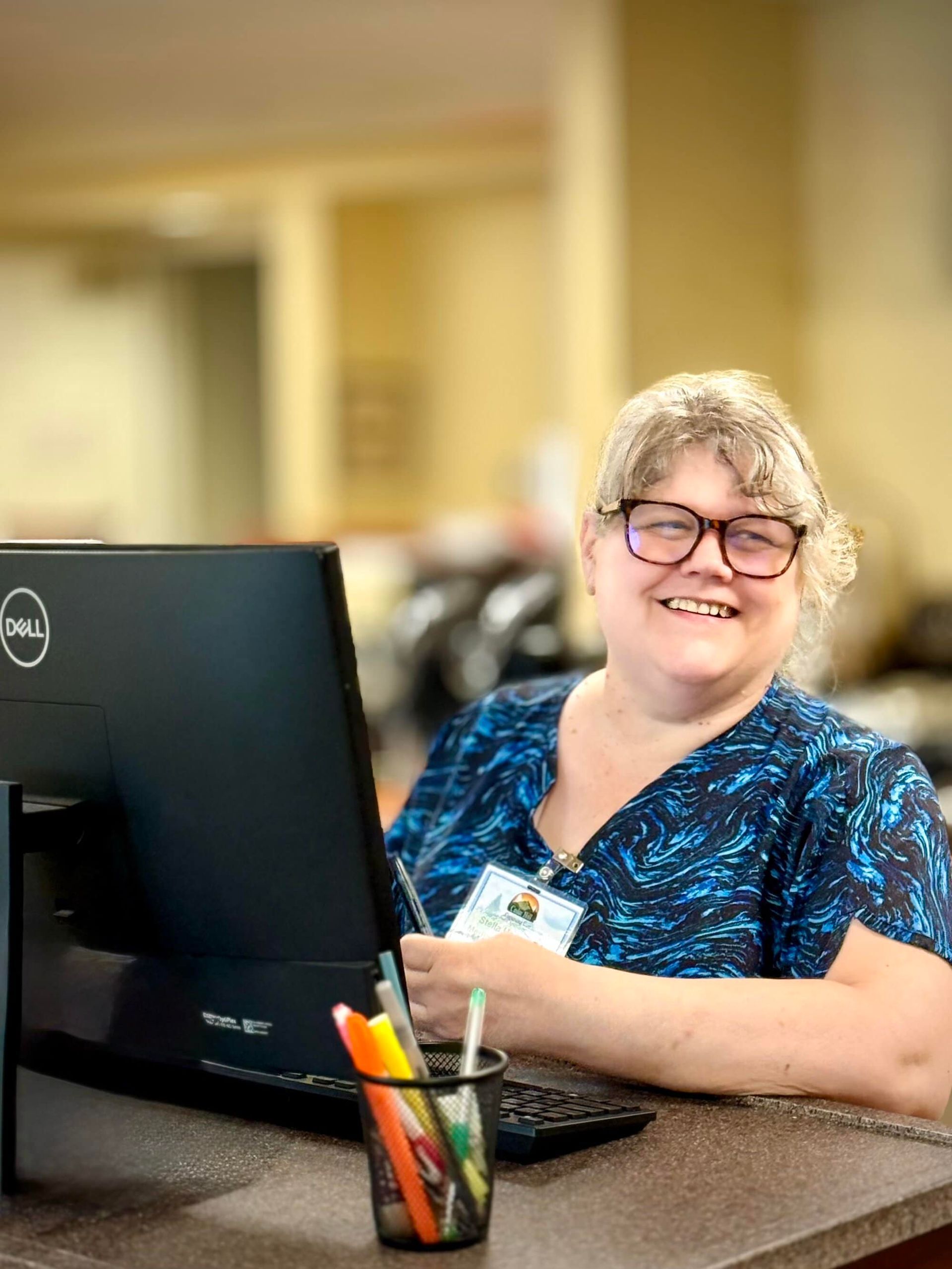 Person smiling at a desk, looking at a Dell computer. Desk holds pens in a cup.