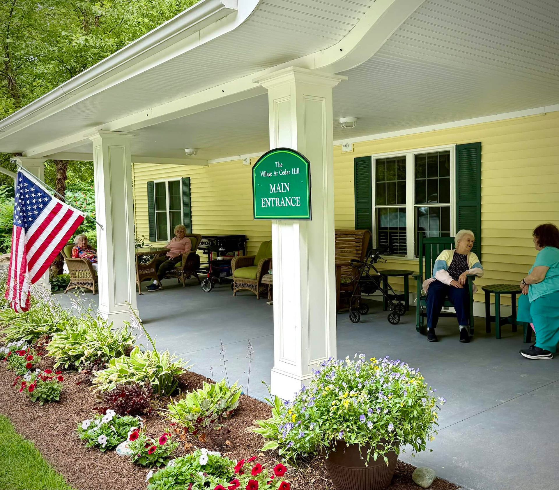 Exterior of assisted living facility with people on porch. American flag, flowerbeds.