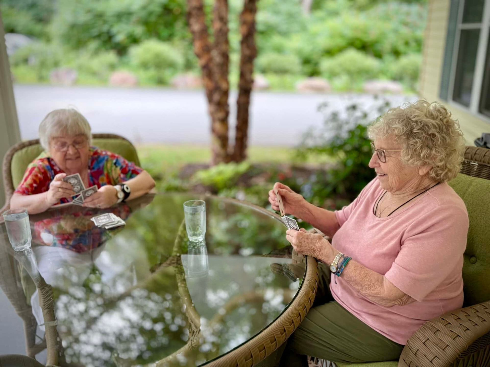 Two older adults play cards at a table outdoors. Both are smiling, with plants visible in the background.