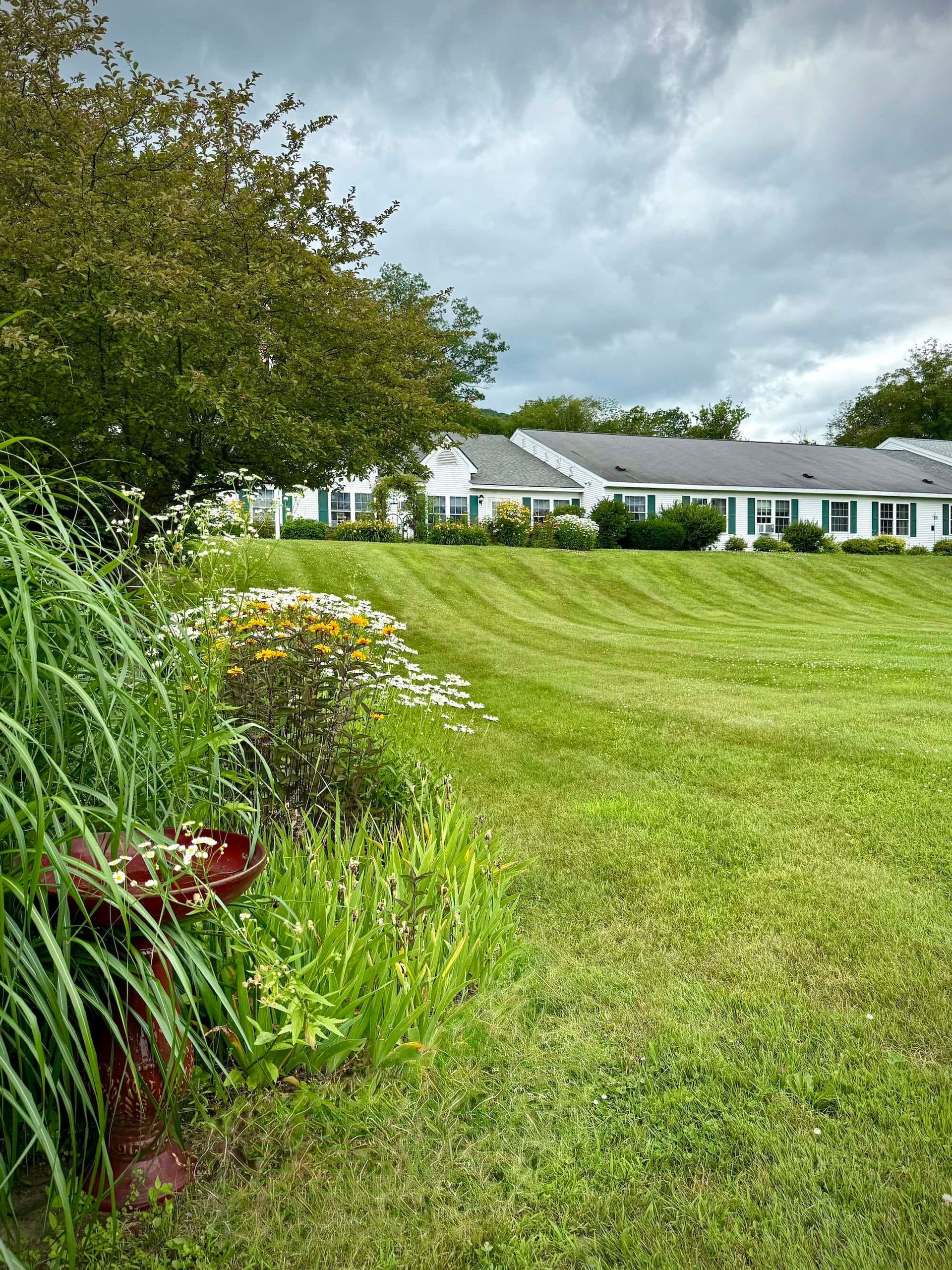 Lawn mowed in stripes slopes toward white buildings under a cloudy sky. A garden with greenery is in the foreground.