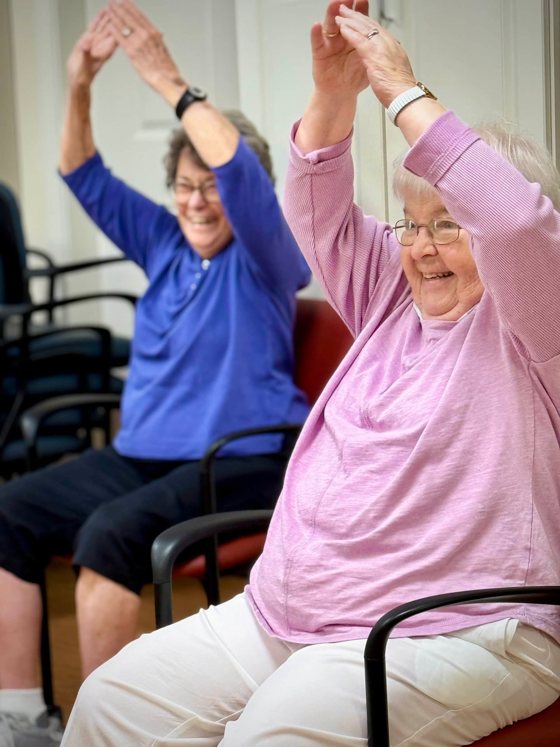 Two women sitting and smiling, stretching arms overhead in a room with chairs.