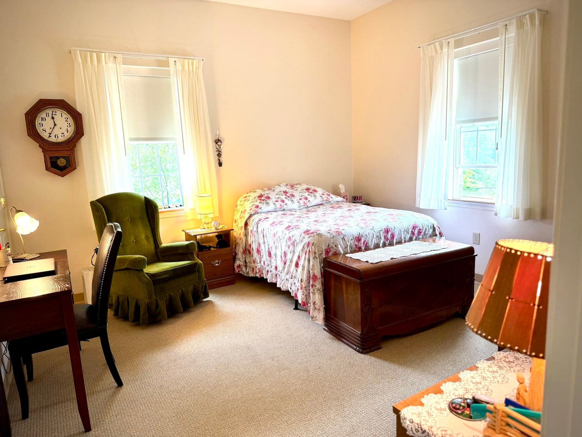 Cozy bedroom with floral bedspread, antique clock, green armchair, and natural light from windows.