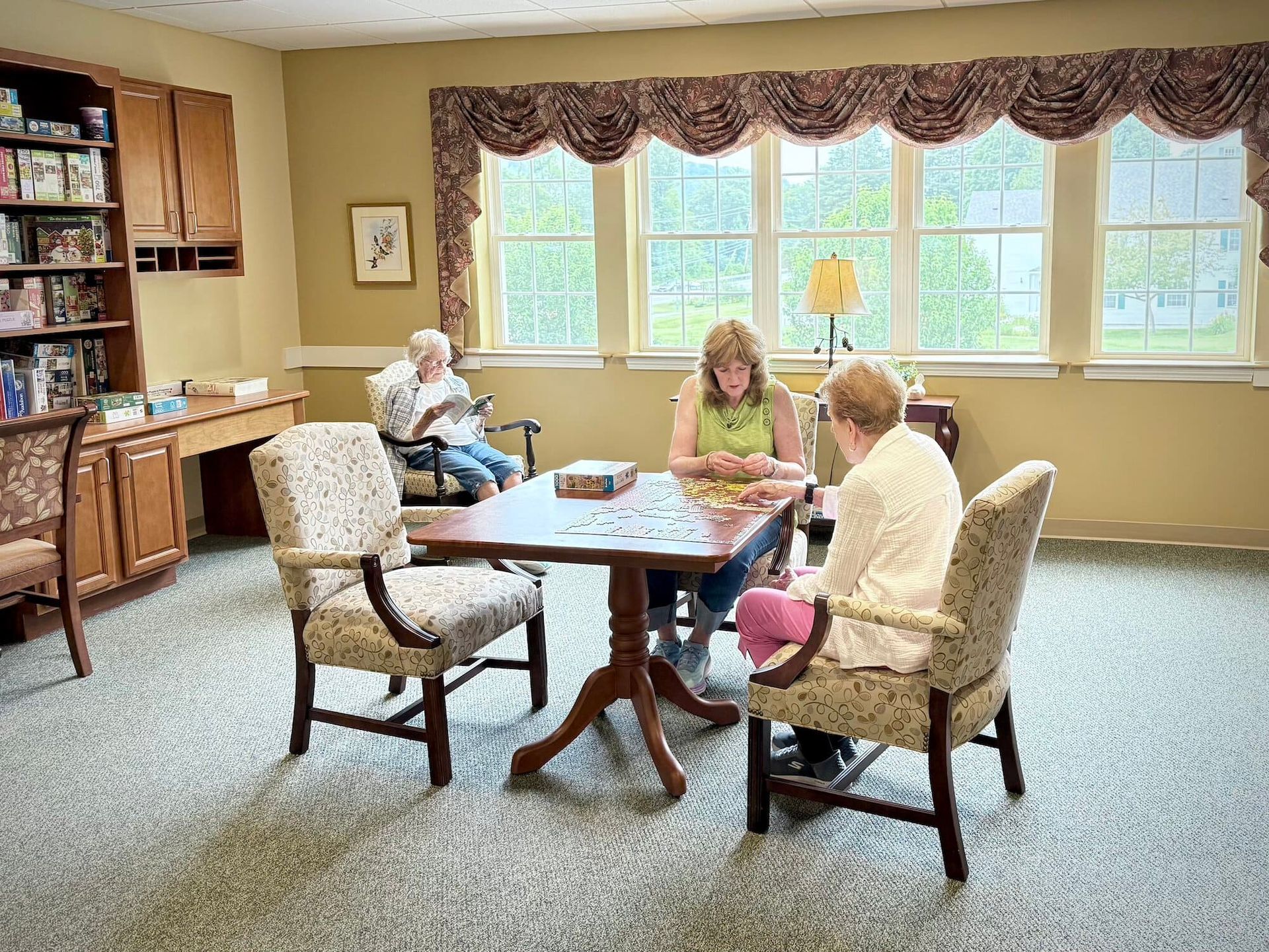 Three people at a table solving a puzzle in a room with a bookcase and window.