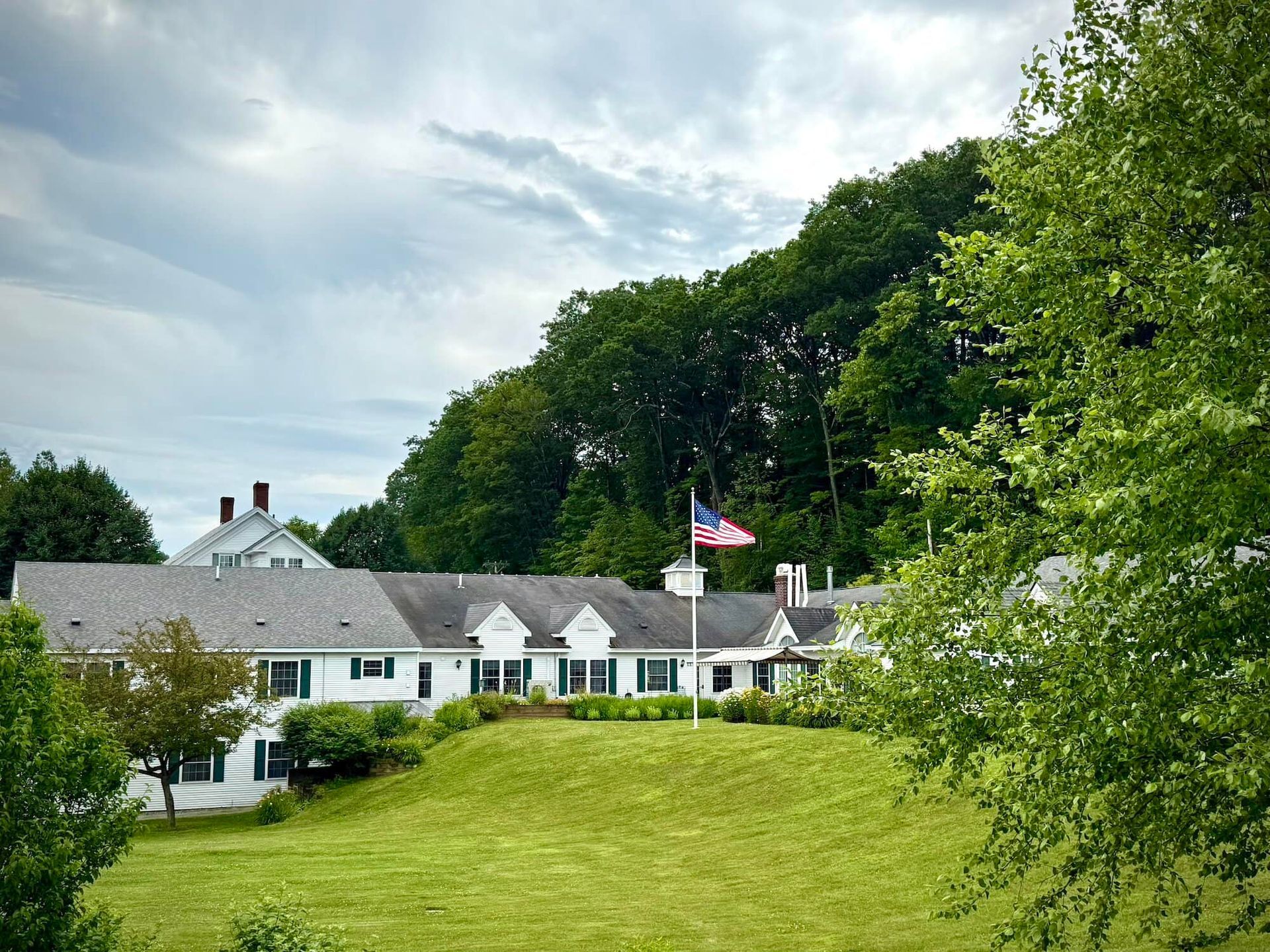 White building with green lawn, trees, and American flag against cloudy sky.