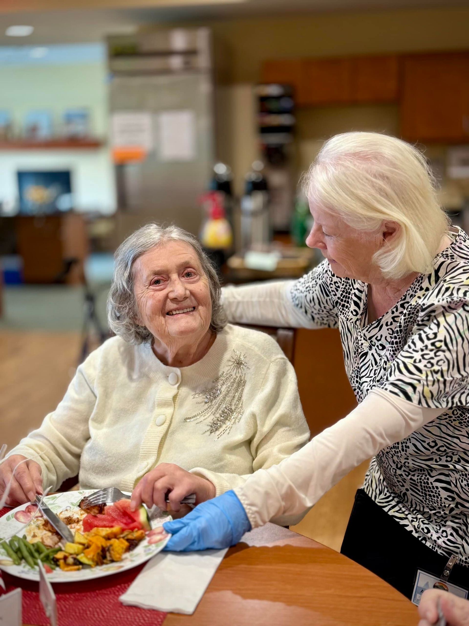 Woman being assisted with a meal at a table by another woman. They are smiling. Indoor setting.