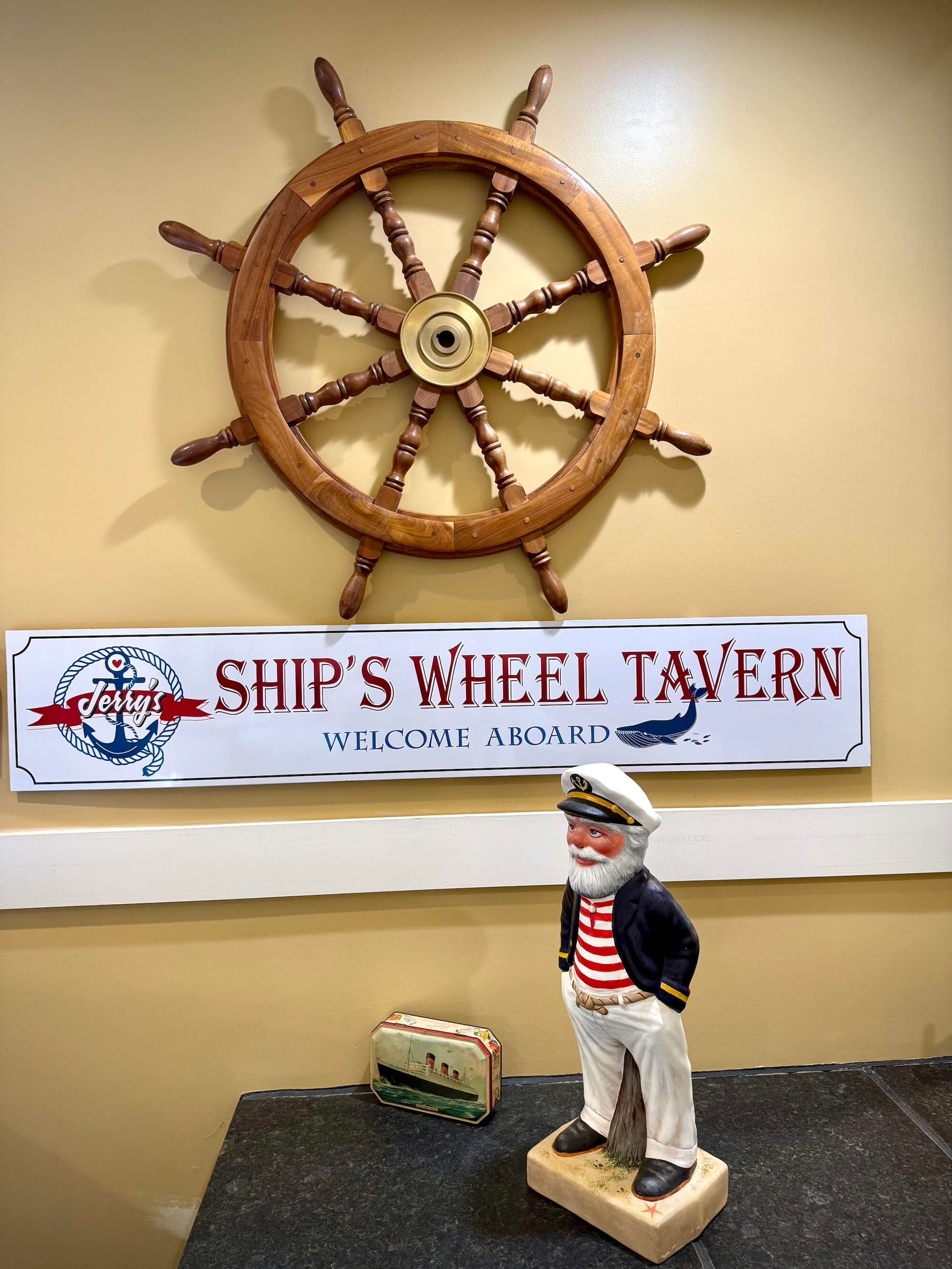 Nautical-themed wall decor with ship's wheel, tavern sign, and sailor figurine on a yellow wall.