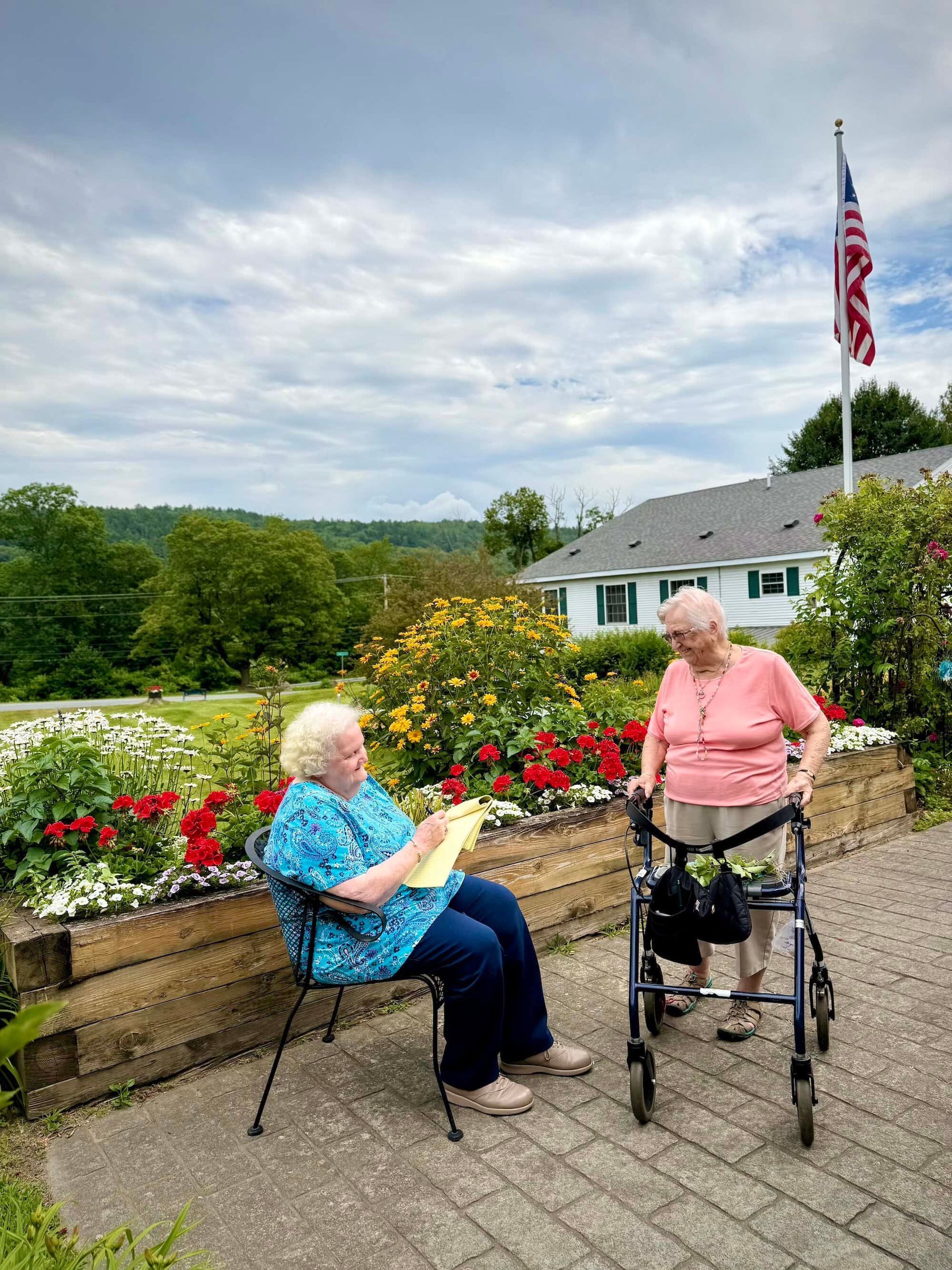 Two women outdoors; one seated drawing, other with walker. Flowers and American flag in background.