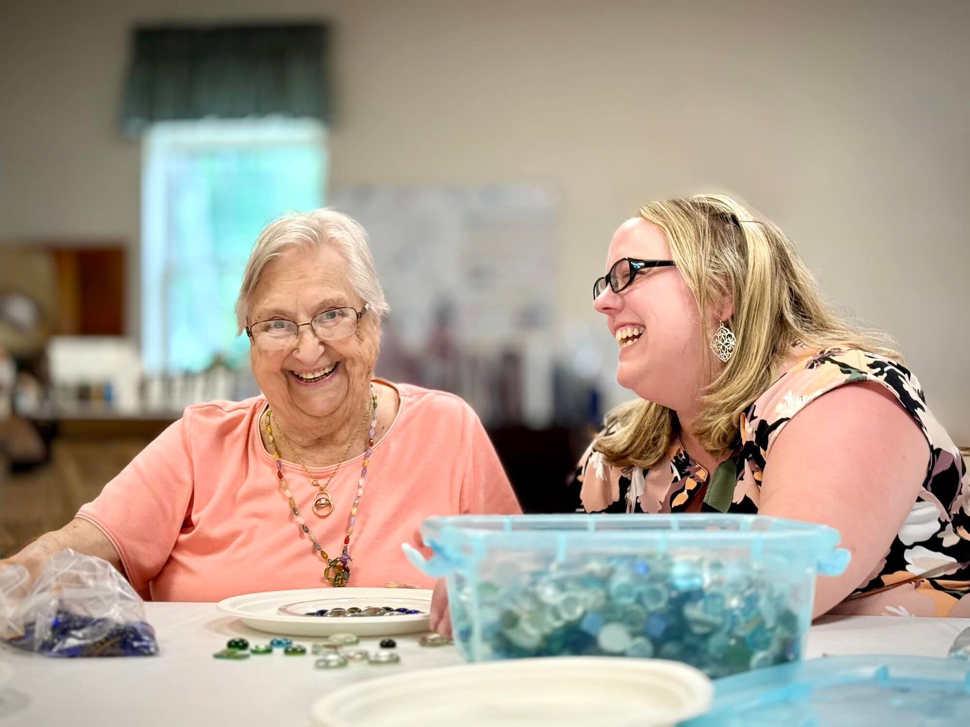 Woman smiles at camera while another laughs, crafting at a table with blue containers and supplies.
