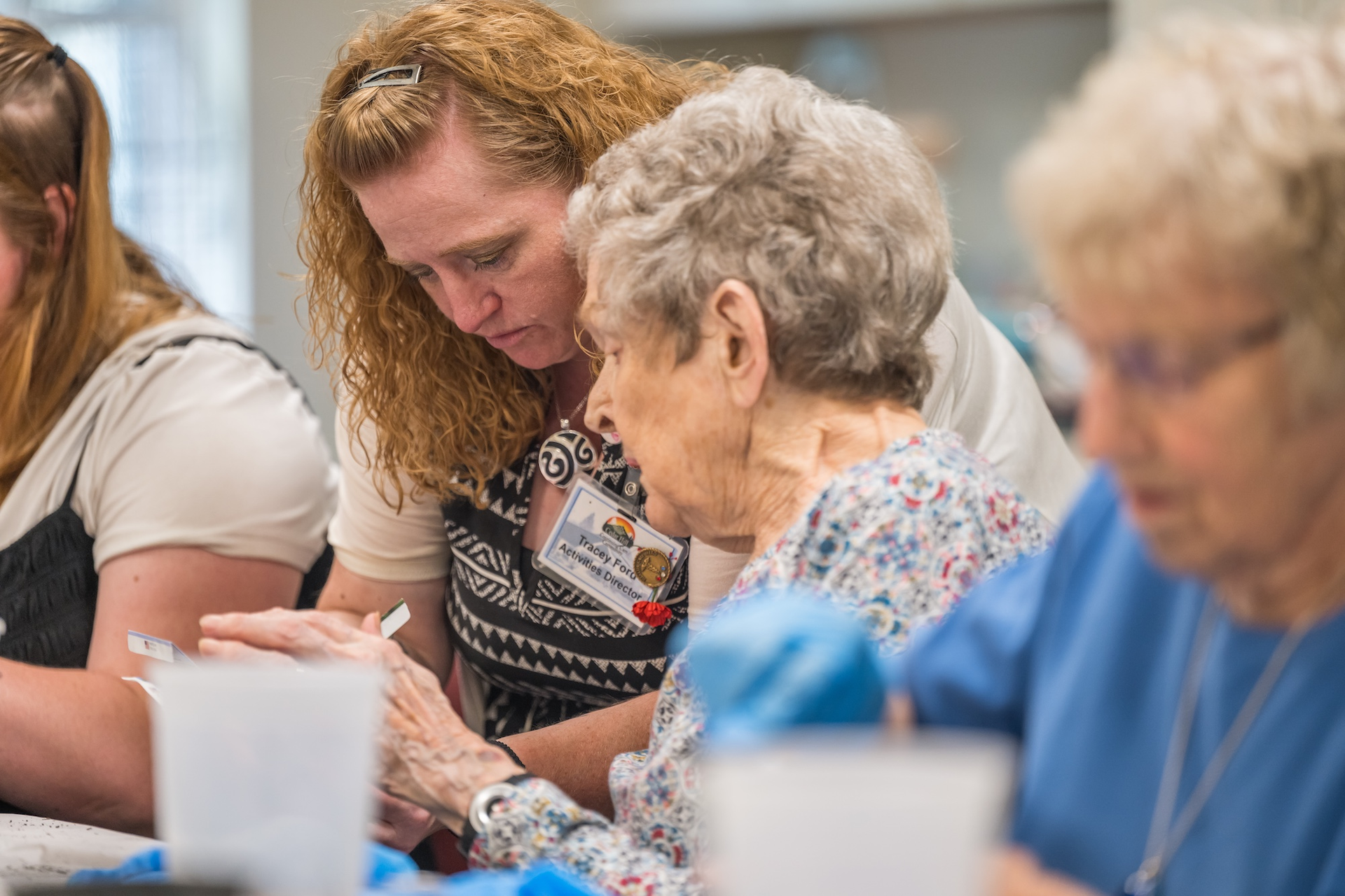 Woman helps elderly person with a task at a table. Others are present, and the setting appears to be indoors.