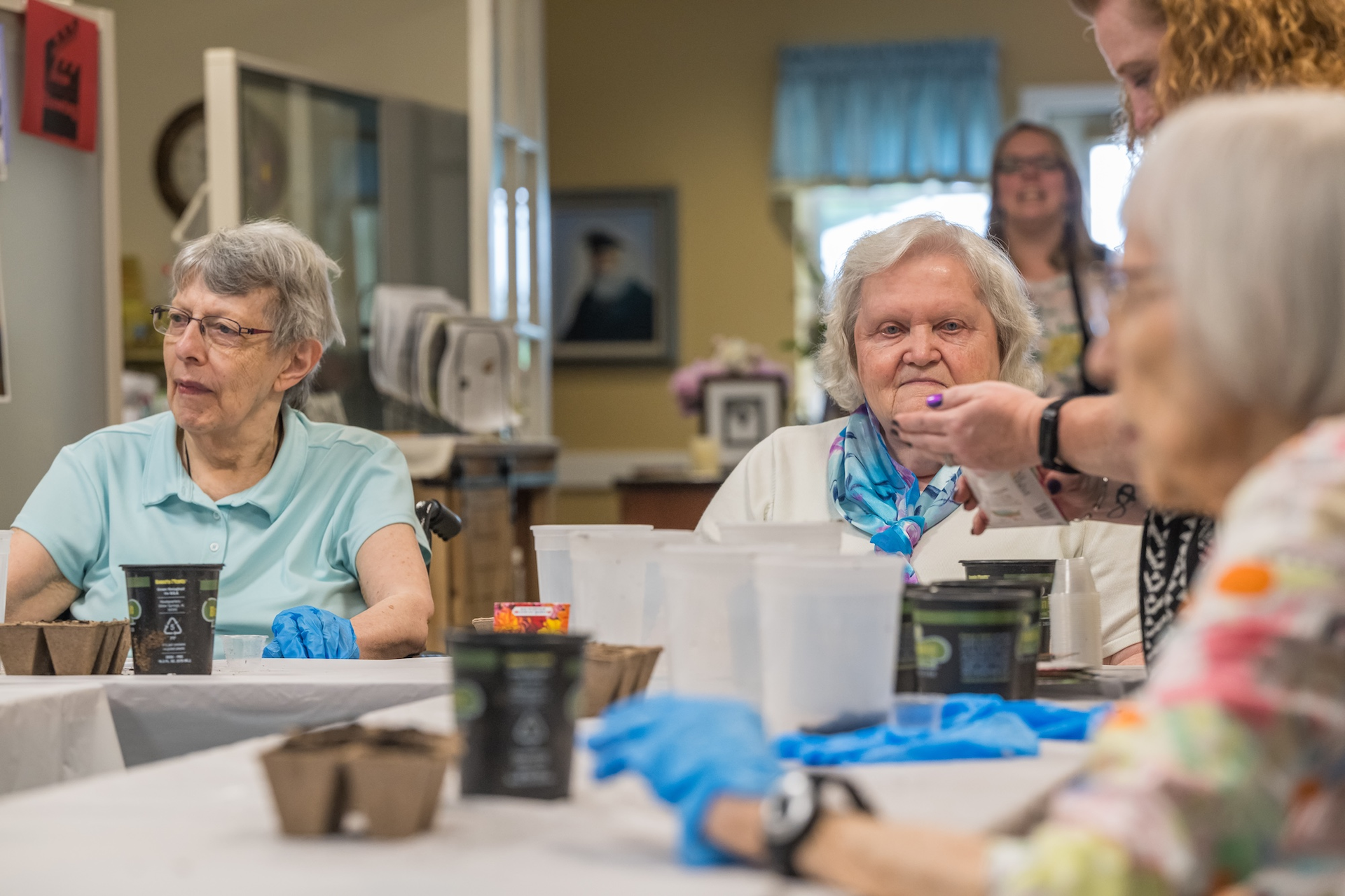 Seniors plant seeds at a table with help from an assistant in a well-lit activity room.
