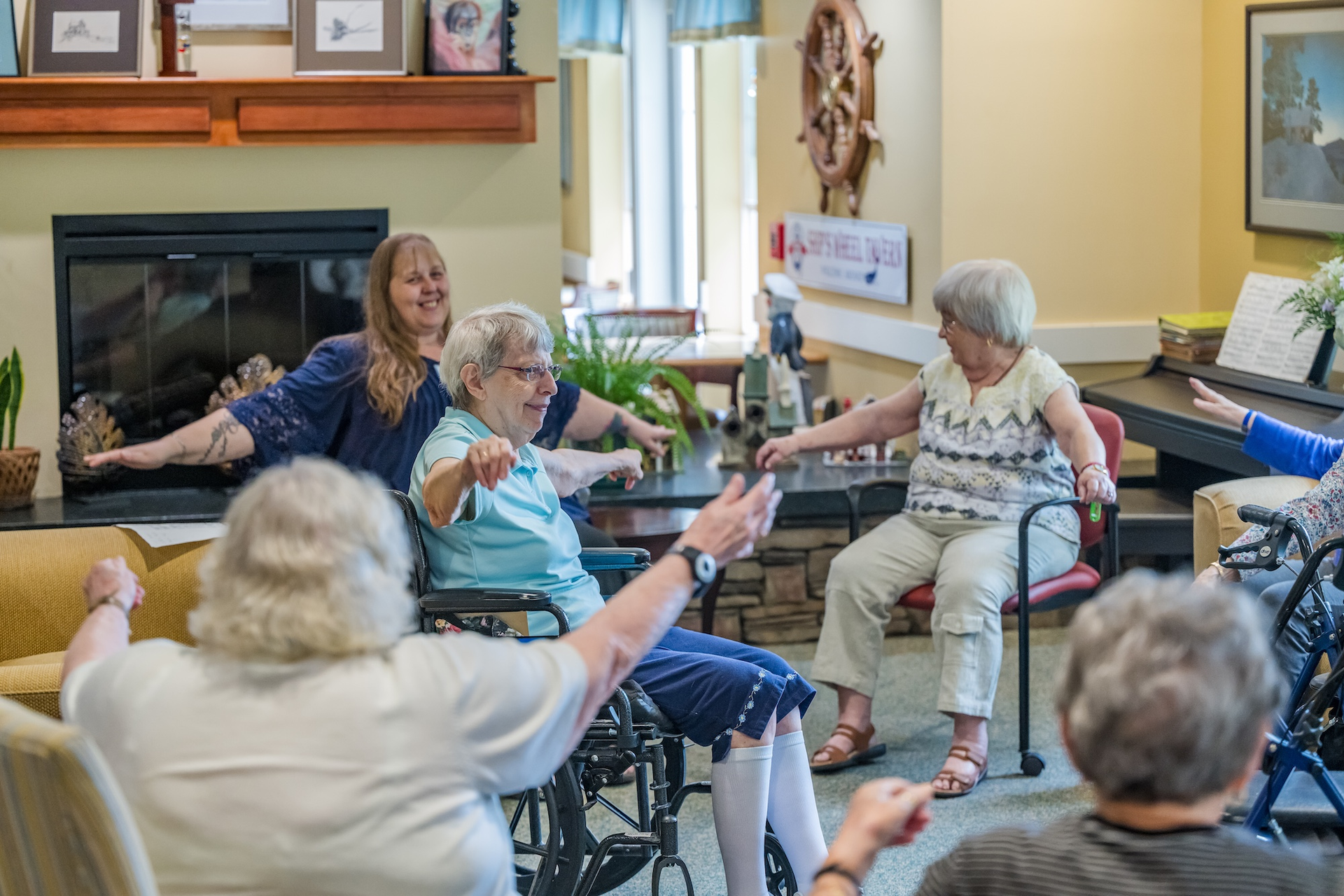 Group of seniors in a room doing seated exercises with a woman. Brightly lit room with fireplace.