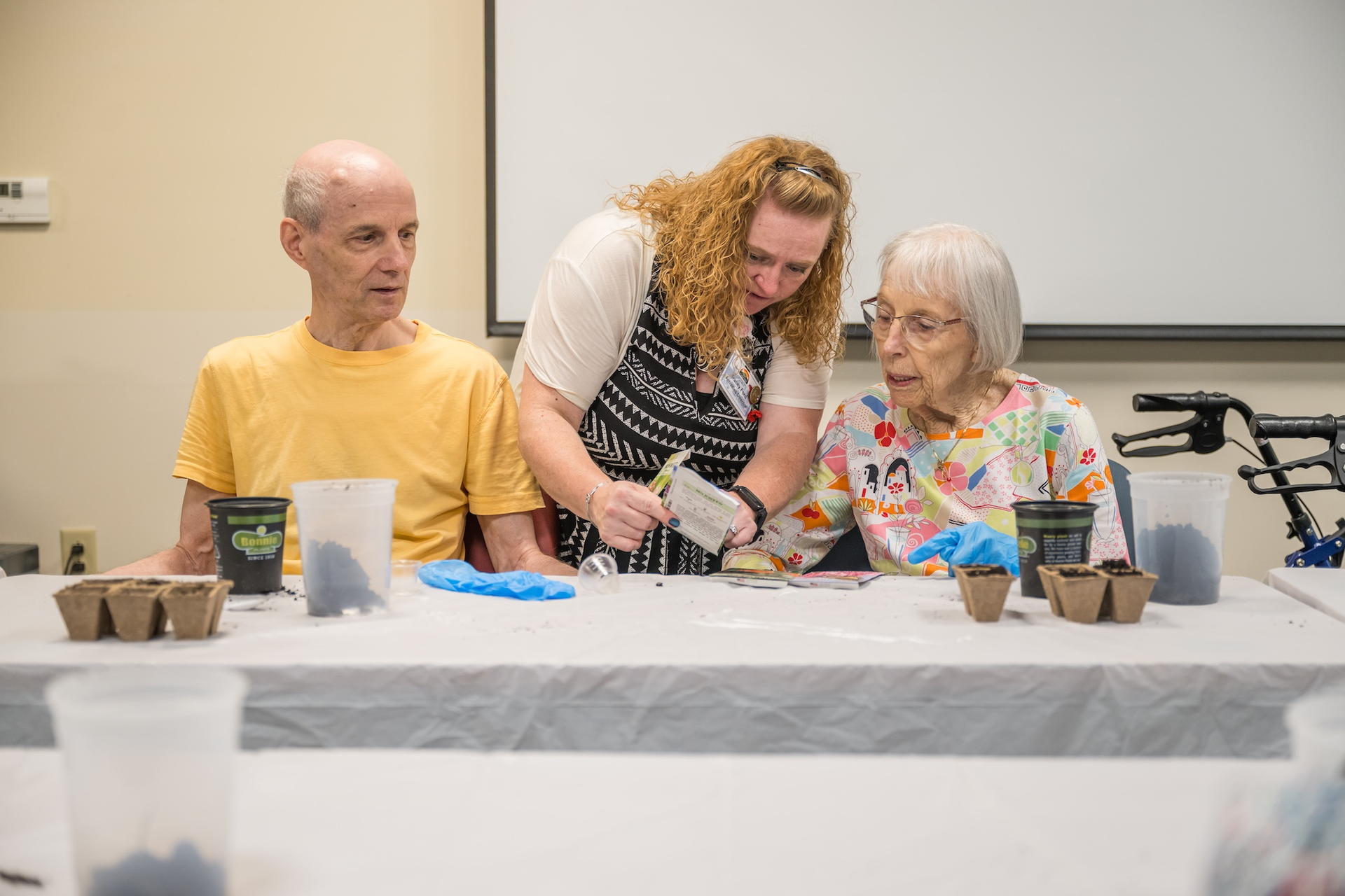 Woman assists two seniors planting seeds at a table, likely in a community center.