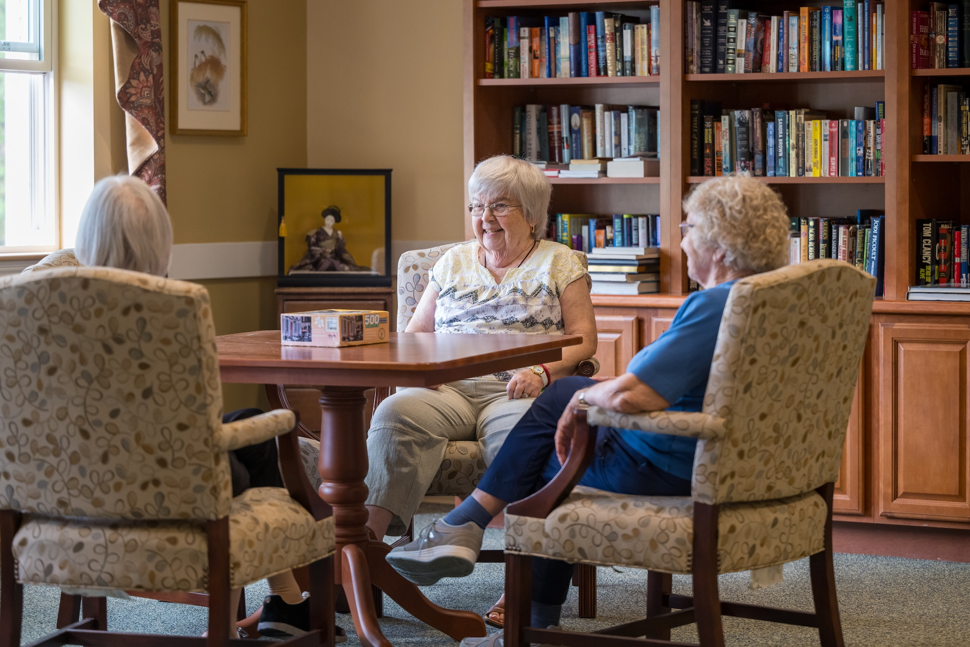 Three older adults seated around a table in a library, talking.