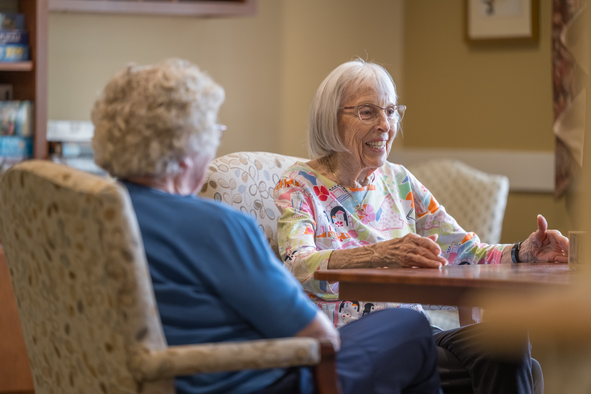 Two older adults sit at a table, conversing. One smiles, gesturing with hands