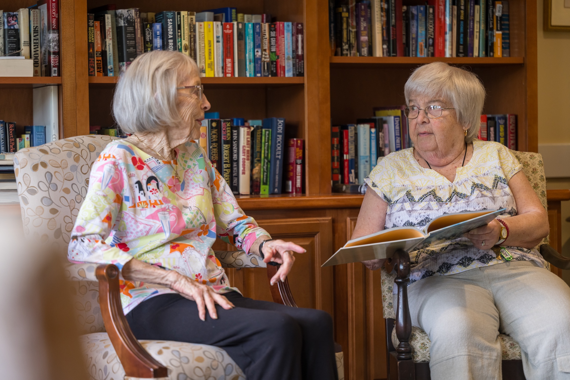 Two women seated in front of a bookshelf, one holding a book, both engaged in conversation.