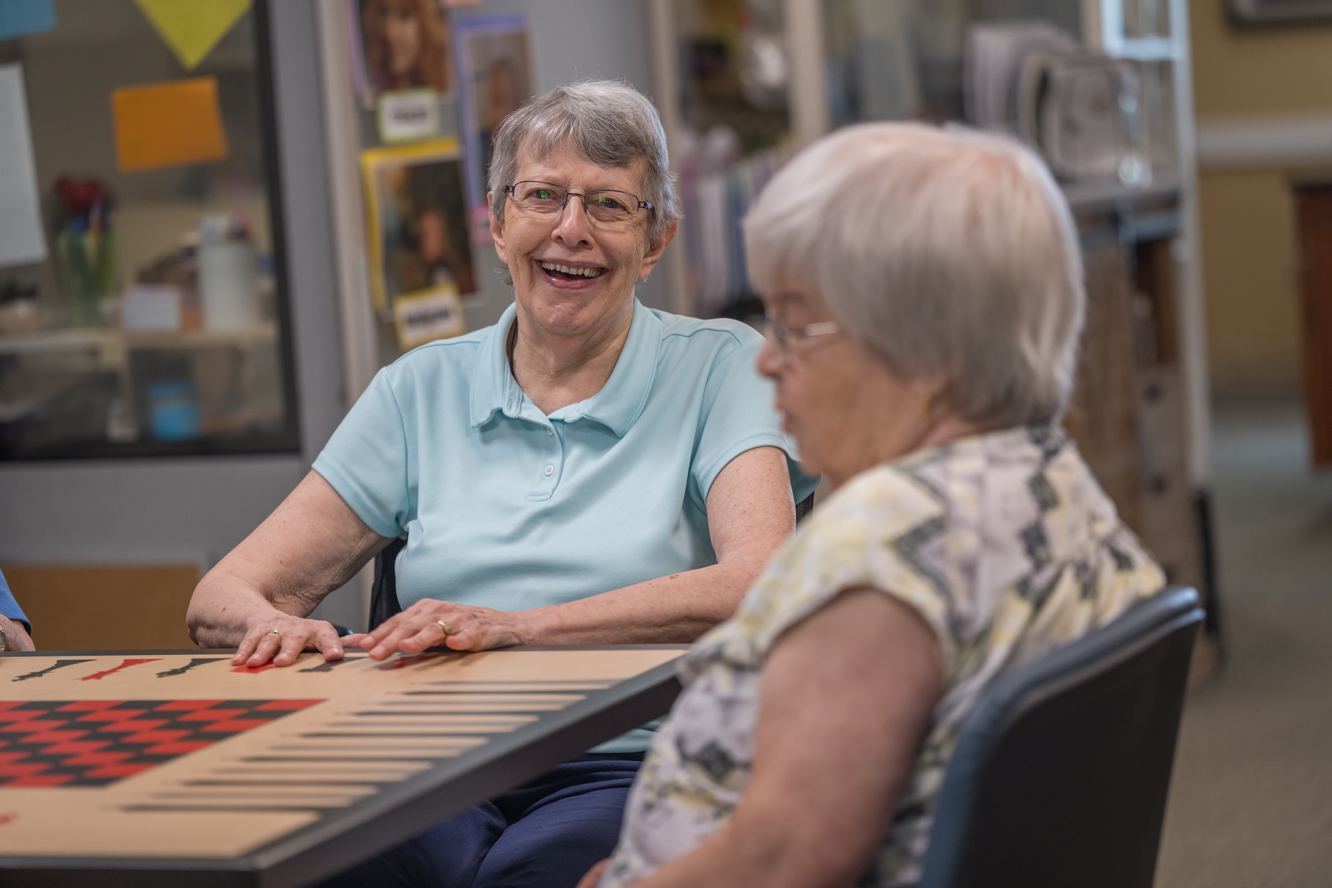 Two women playing a board game at a table, one smiling, in a brightly lit room.