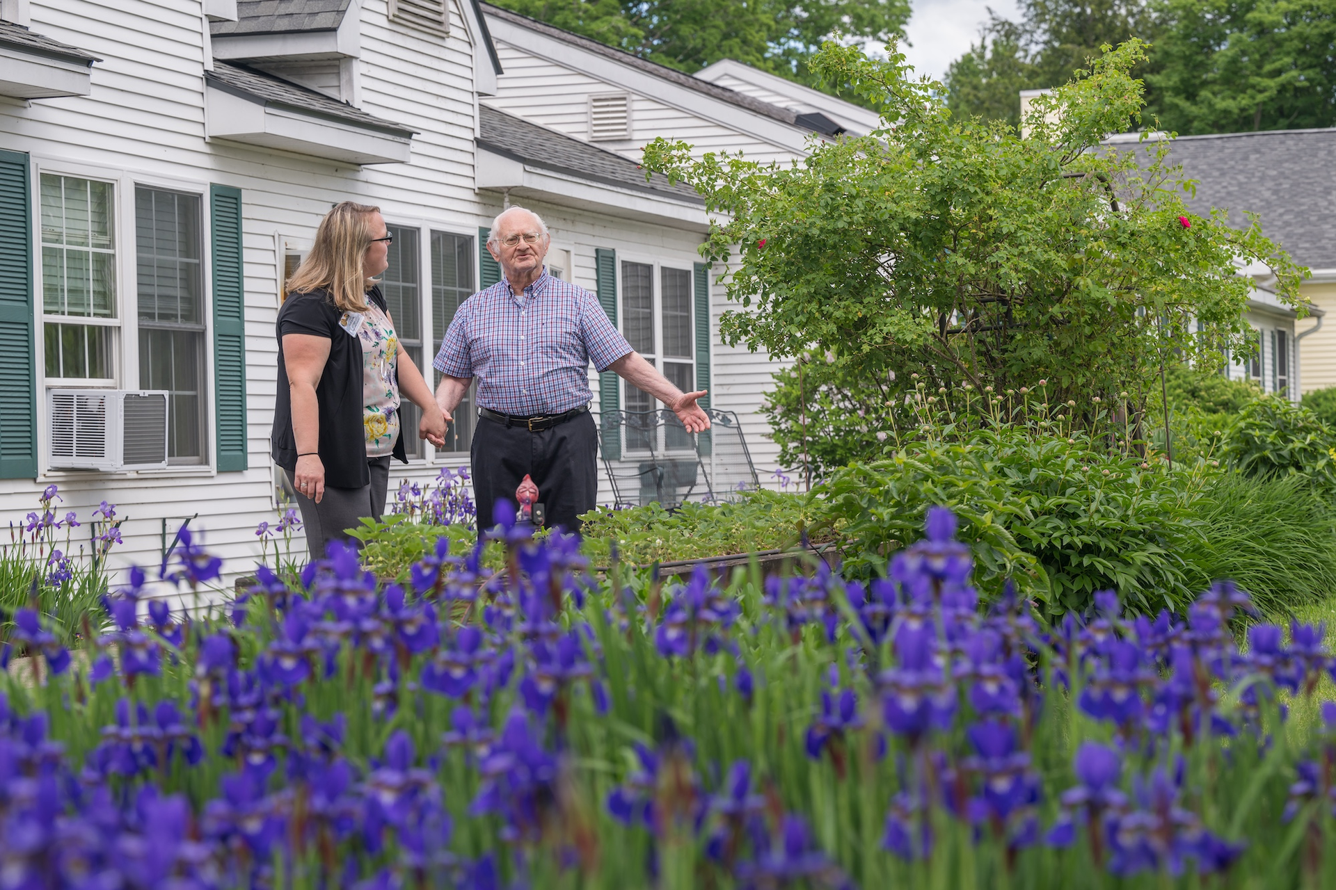 Man and woman standing in front of white house, surrounded by purple iris flowers. Man gestures toward the garden.