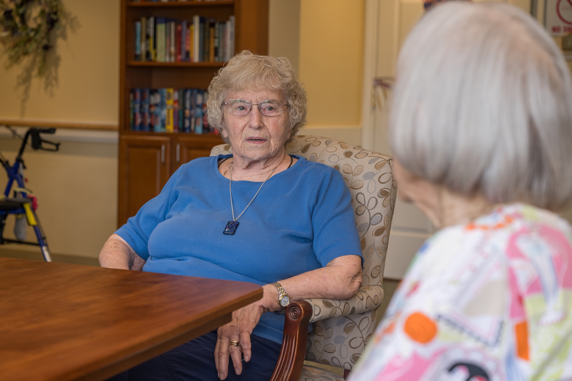 An older woman in a blue shirt sits at a table, facing another person.  A bookshelf and a walker are in the background.