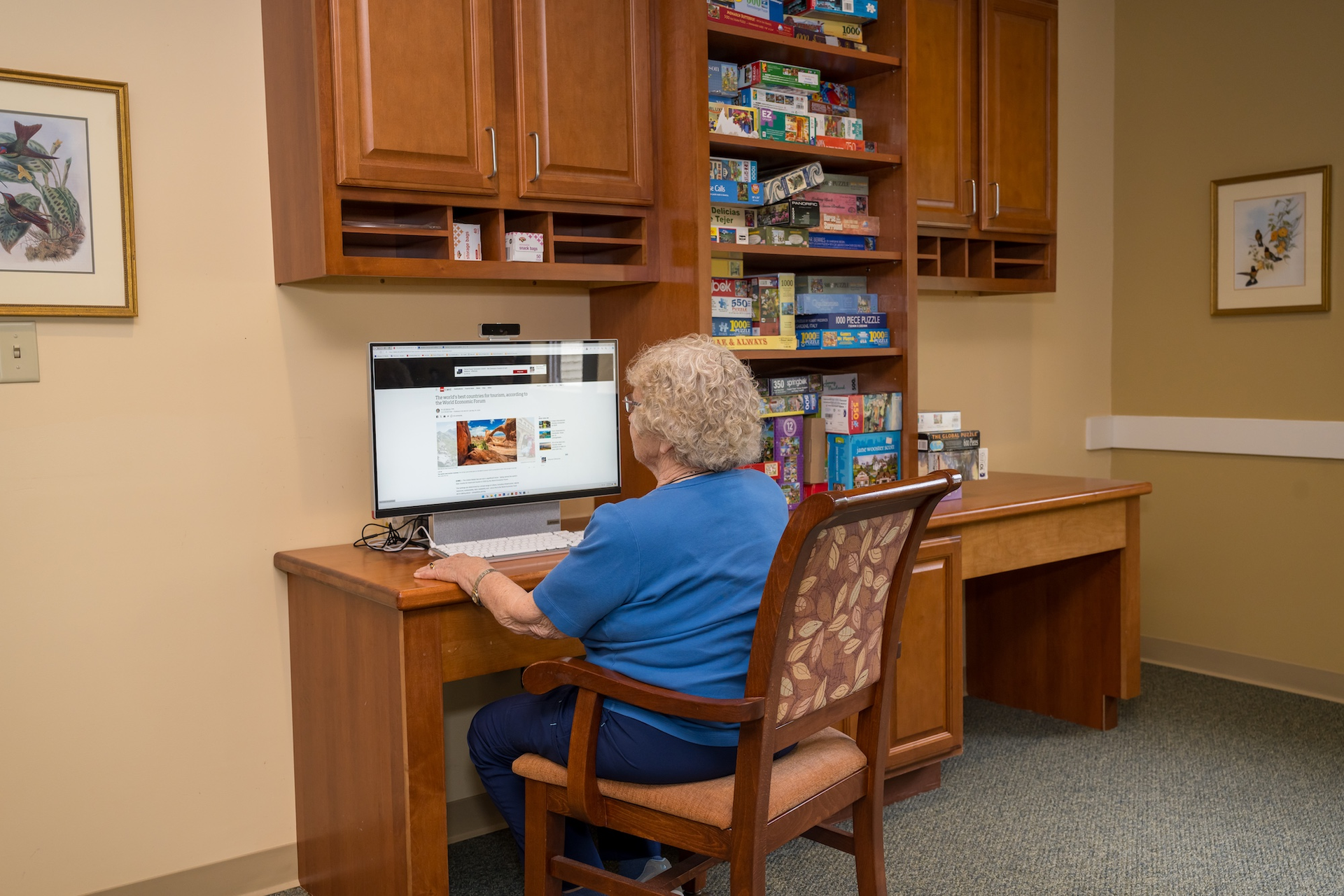 Woman sitting at a desk, using a computer. Wooden cabinets and shelves filled with items are behind her.