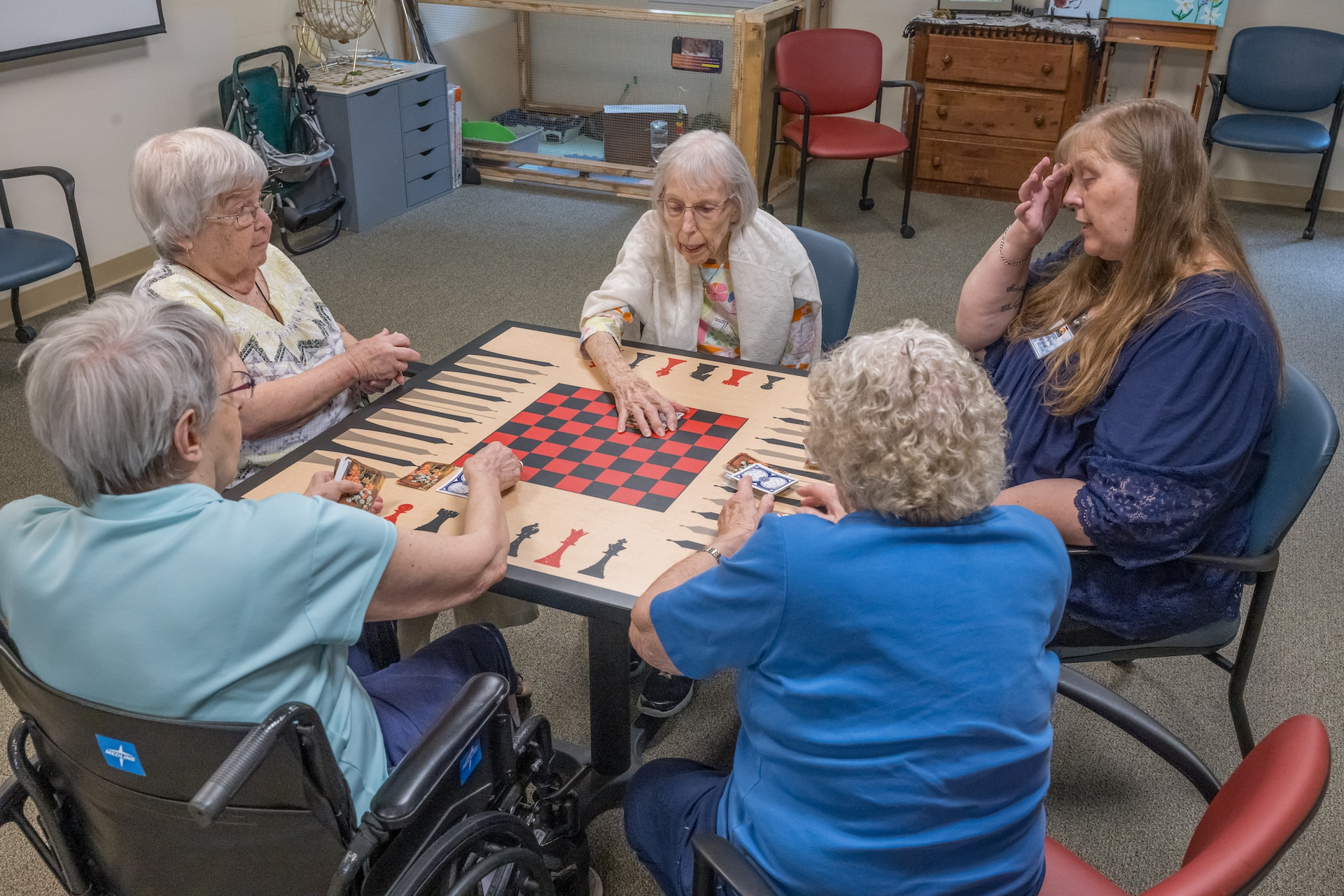 Five people playing a board game at a table. One is in a wheelchair. One woman is touching her head.