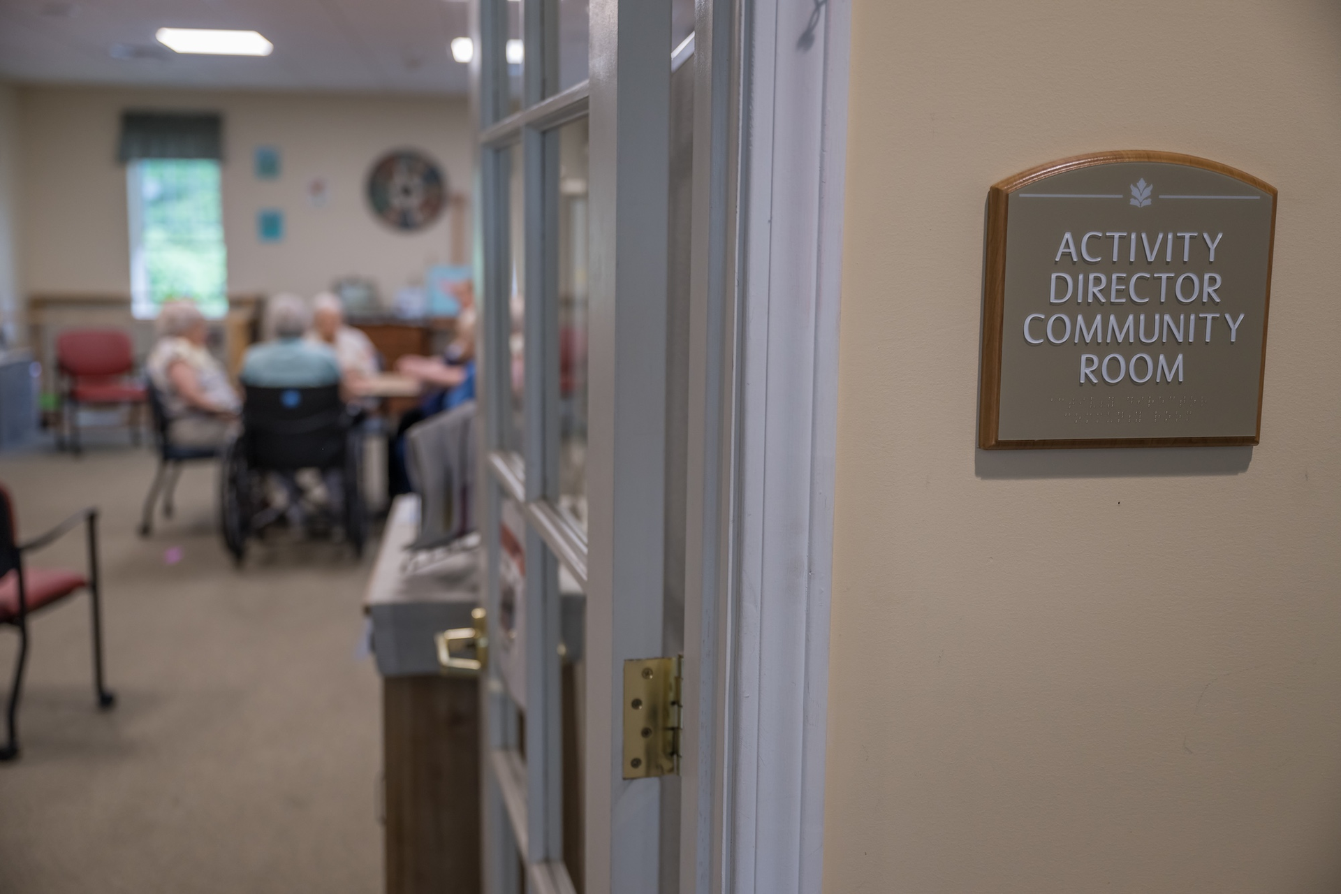 Doorway to Activity Director Community Room with sign. Elderly people seated at a table, in the background.