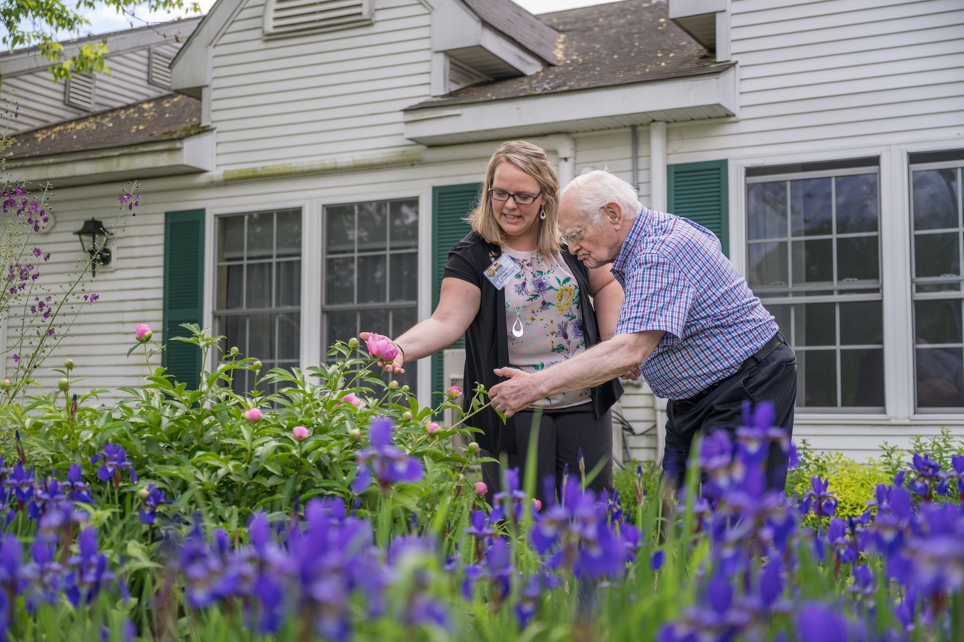 Woman and older man looking at flowers in a garden. White house in background.