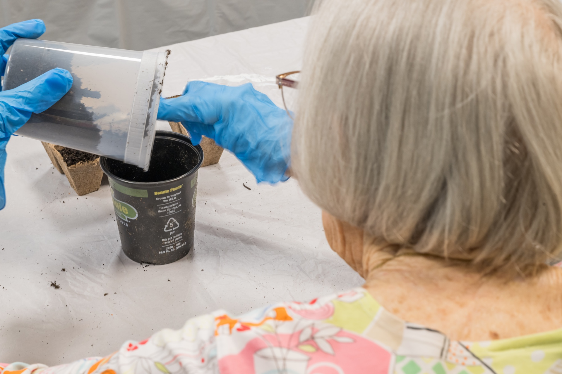 Hands in blue gloves pouring soil into a black plant pot, near a person with gray hair.