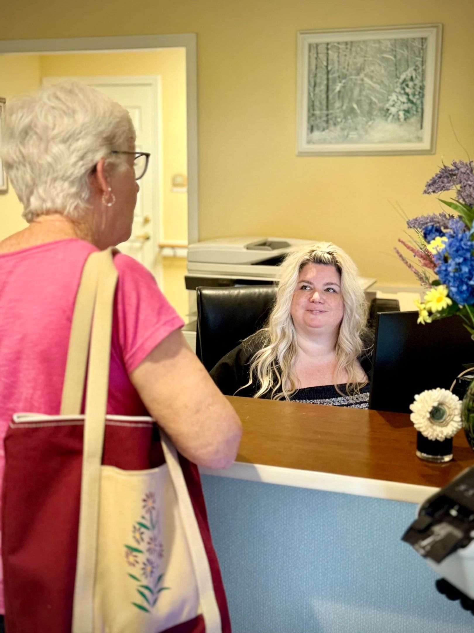 Woman with a tote bag at a reception desk, talking to a person seated behind it. Light-colored walls, flowers.