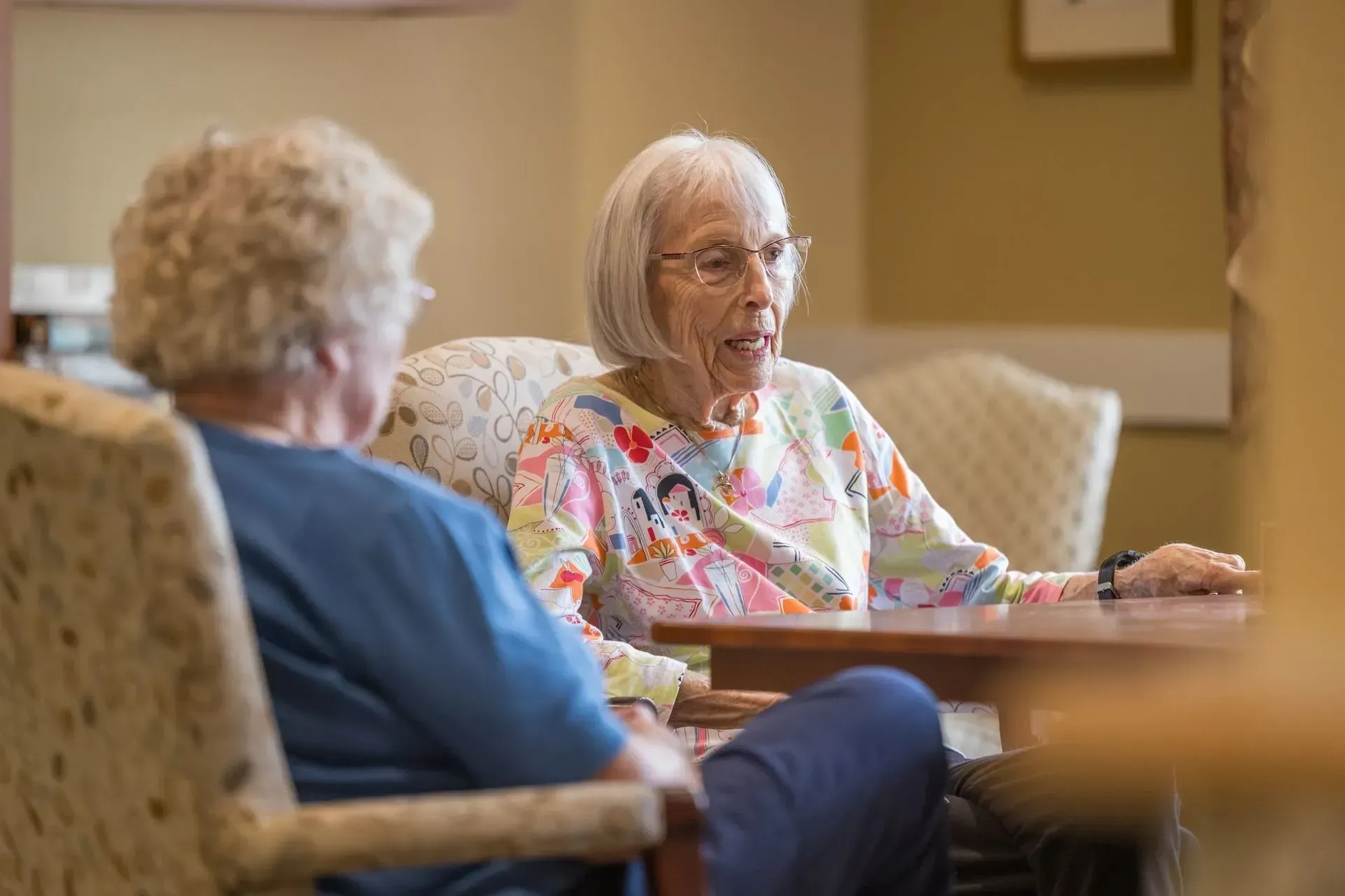 Two older women seated at a table, conversing indoors. One wears glasses, a patterned shirt. Beige walls.