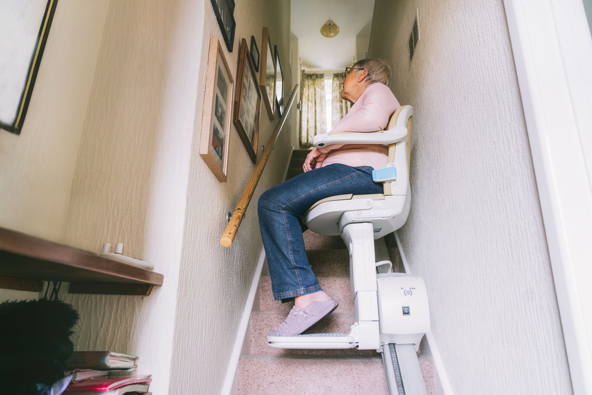 A person wearing light-colored clothing and dark trousers sits on a stairlift moving upward along a residential staircase.