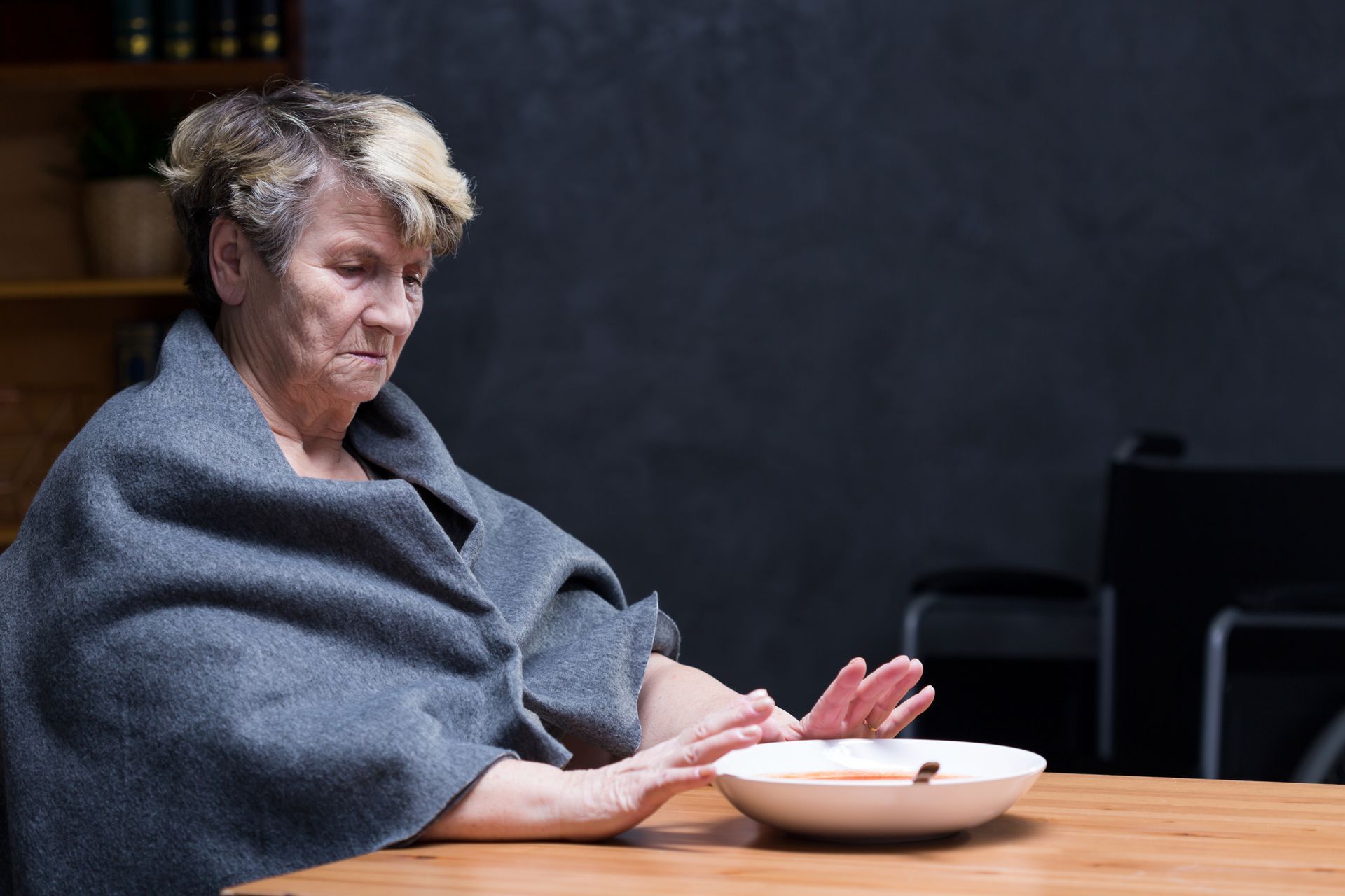 Older person at a table, refusing food in a bowl. Wrapped in a shawl, somber expression, wheelchair in the background.