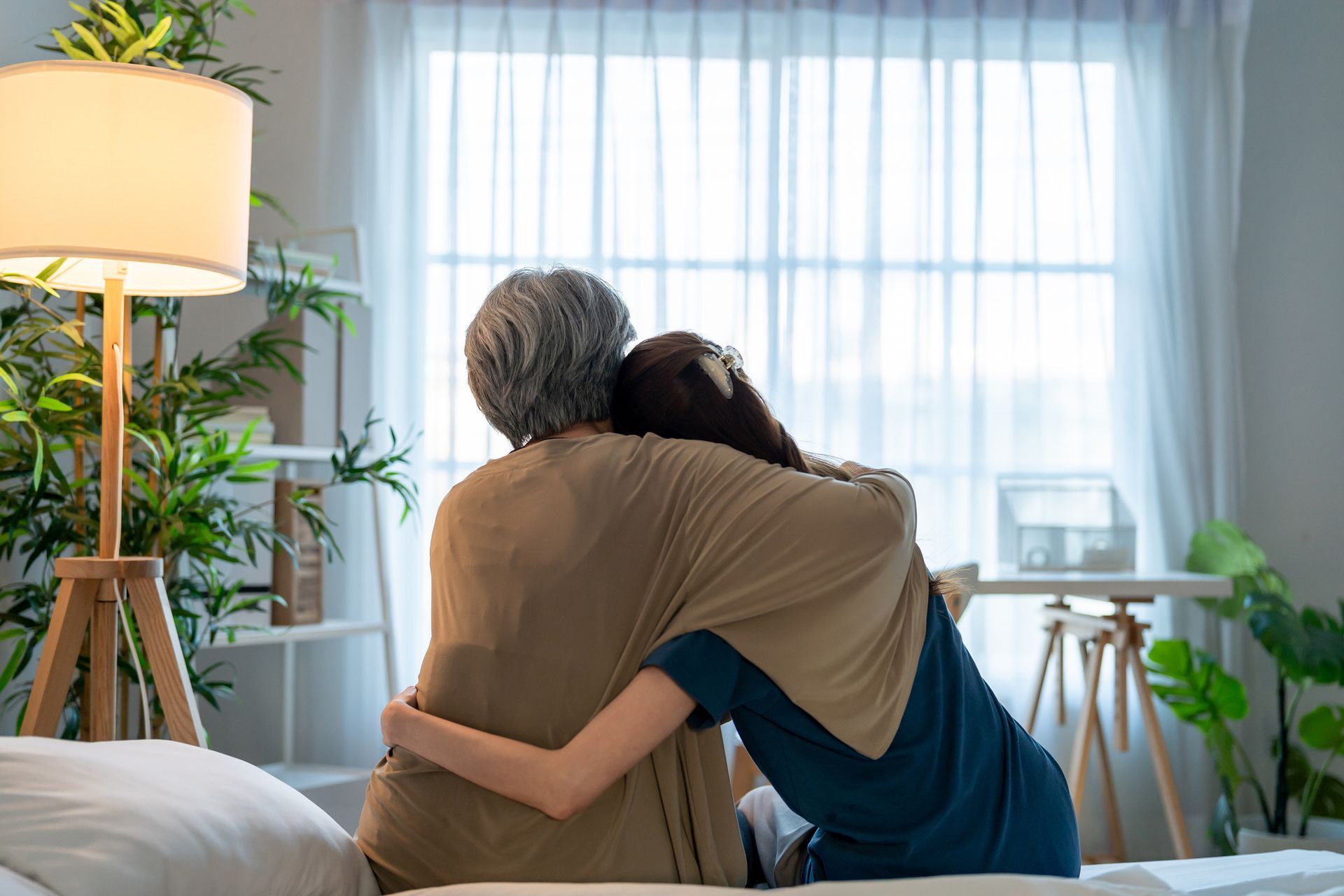 Woman hugging another person on a bed, looking out a window. Beige shawl, bright room.