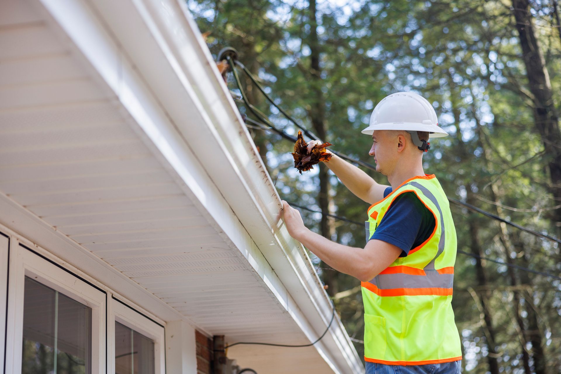 A worker in a high-visibility vest and hard hat installing or repairing a black cable along a white house gutter.