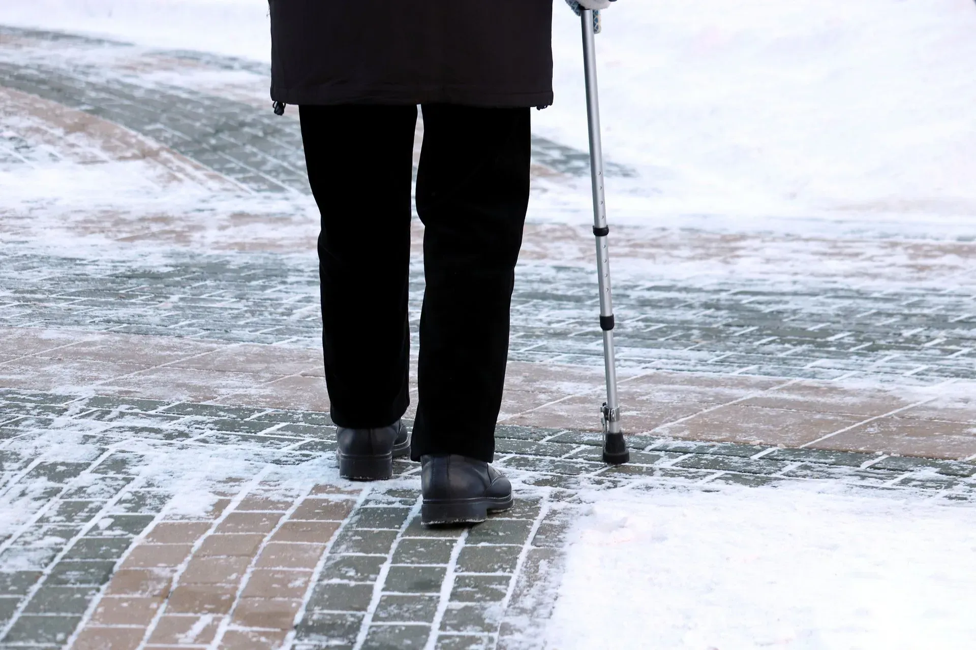 Person walking with a cane on a snow-covered brick path.