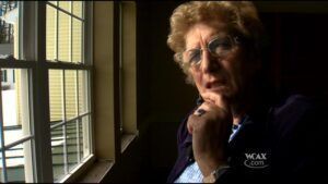 Woman with glasses, hand on chin, looking pensively near a window.