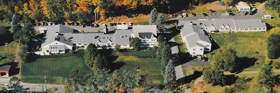 Aerial view of a light-colored building with multiple sections and a large lawn surrounded by trees.