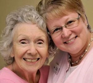 Two smiling women, one elderly with gray hair, the other middle-aged with glasses, both wearing pink.
